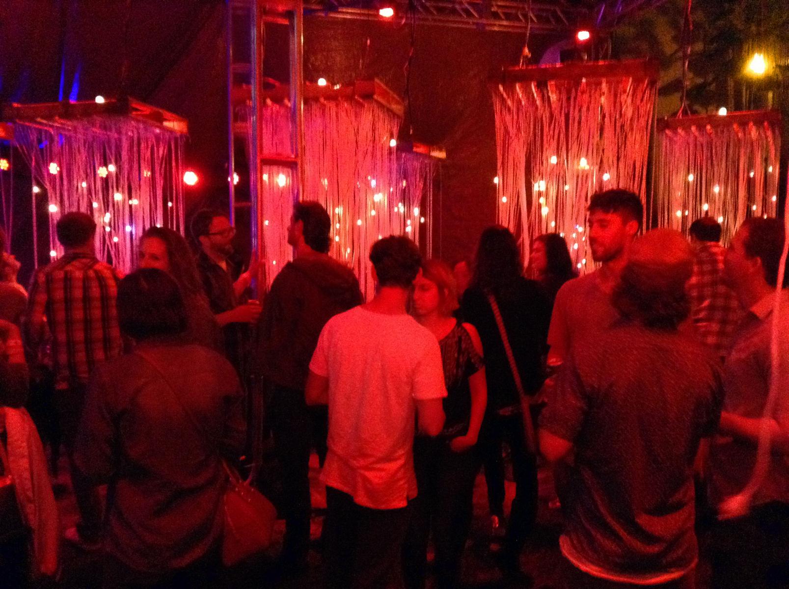 Ipomoea:Grass, bathed in red fire-like light, strings and points of light hand from suspended shipping palettes around dozens of visitors.